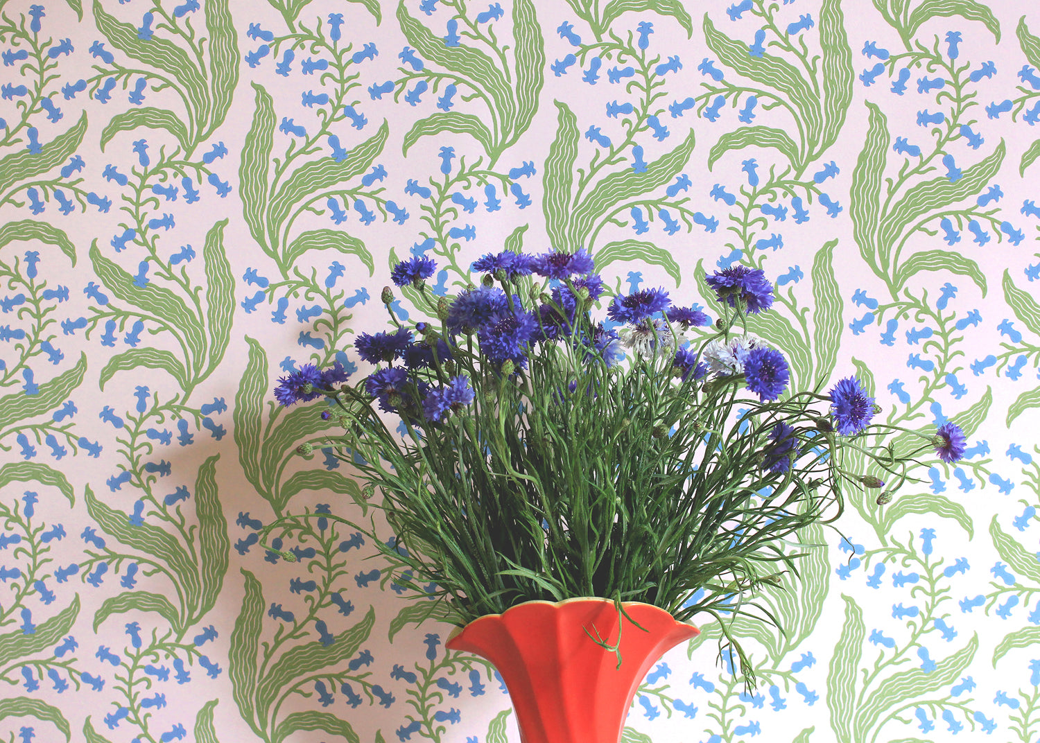 Red vase and flowers against wallpaper in a scrolling floral pattern in blue and green on a pink field. 