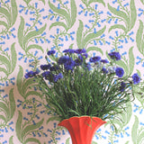 Red vase and flowers against wallpaper in a scrolling floral pattern in blue and green on a pink field. 