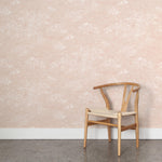 A wooden chair stands in front of a wall papered in a painterly cloud pattern in white on a pastel pink field.