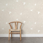 A wooden chair stands in front of a wall papered in a print of birds and stars in white on a soft beige watercolor field.
