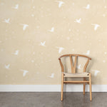 A wooden chair stands in front of a wall papered in a print of birds and stars in white on a pastel yellow field.