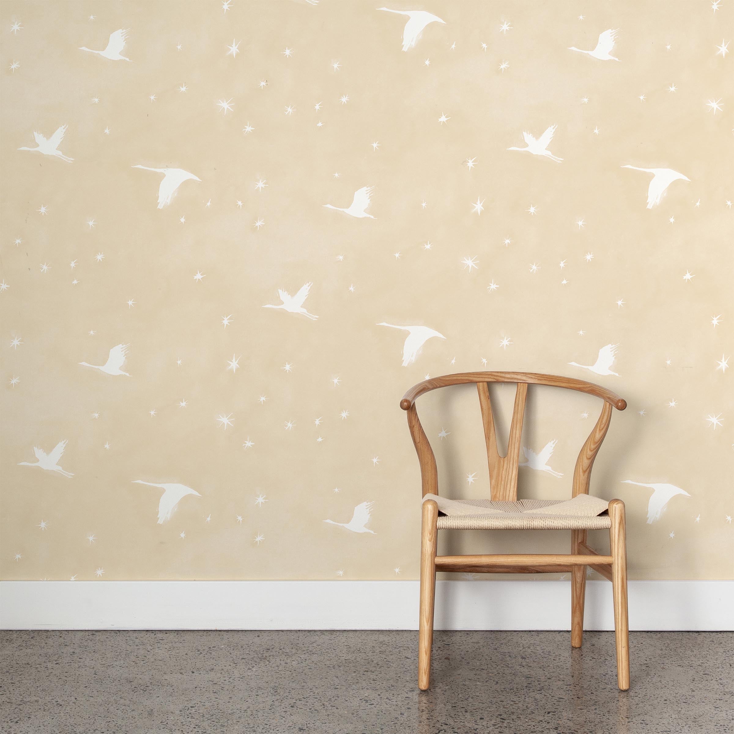 A wooden chair stands in front of a wall papered in a print of birds and stars in white on a pastel yellow field.