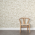 A wooden chair stands in front of a wall papered in a painterly branch print in white on a taupe watercolor field.