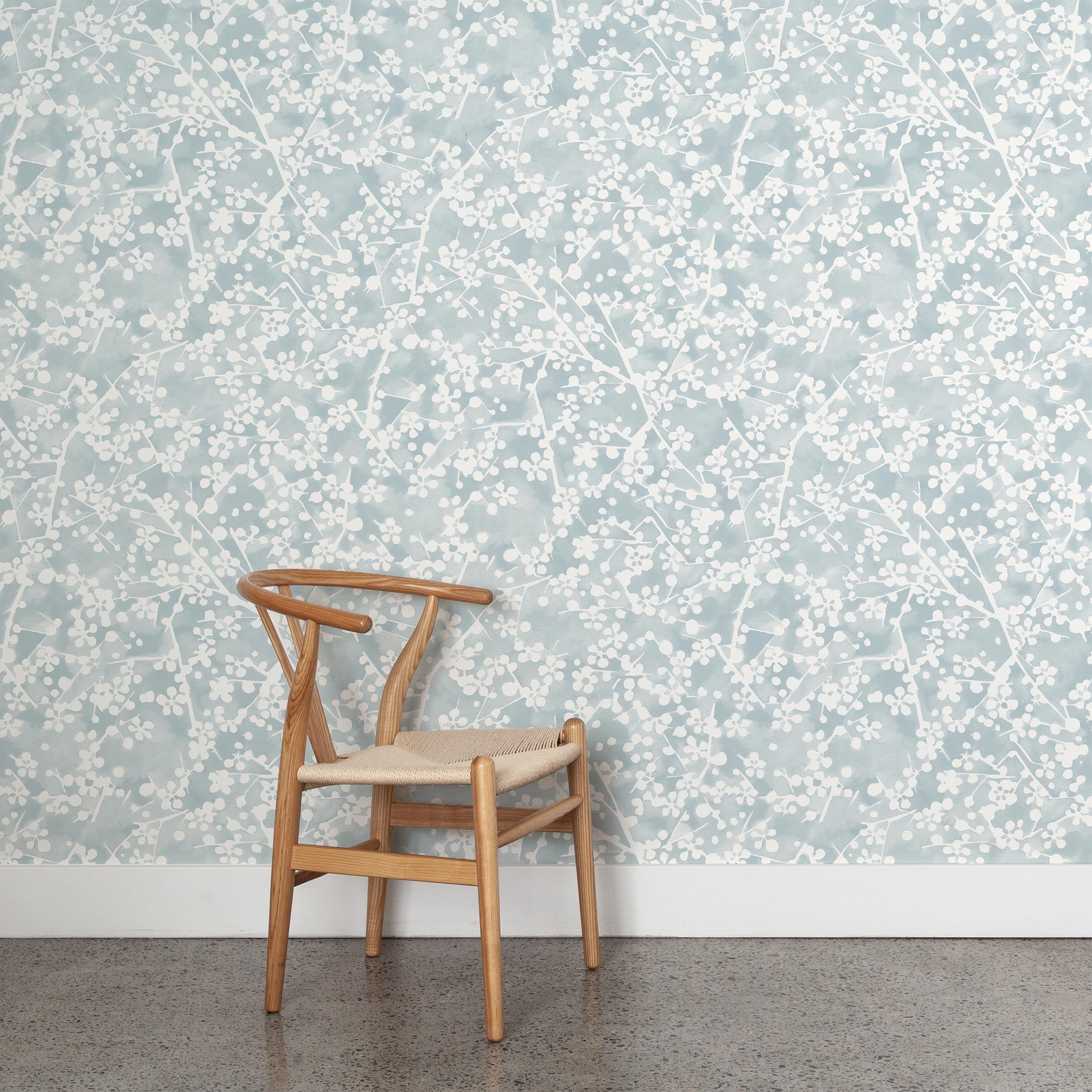 A wooden chair stands in front of a wall papered in a painterly branch print in white on a warm blue watercolor field.