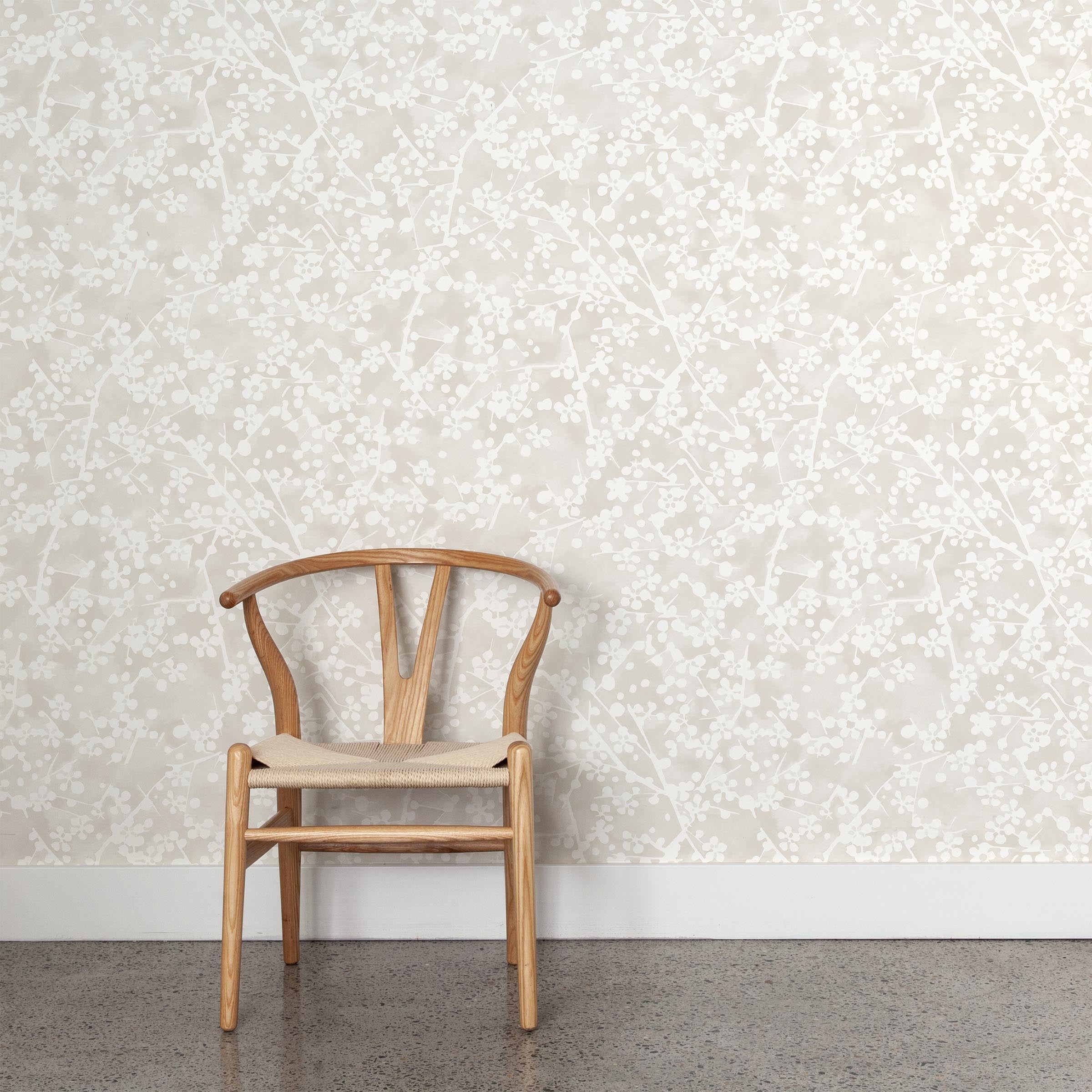 A wooden chair stands in front of a wall papered in a painterly branch print in white on a soft beige watercolor field.
