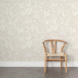 A wooden chair stands in front of a wall papered in an abstract leaf pattern in white on a soft beige watercolor field.