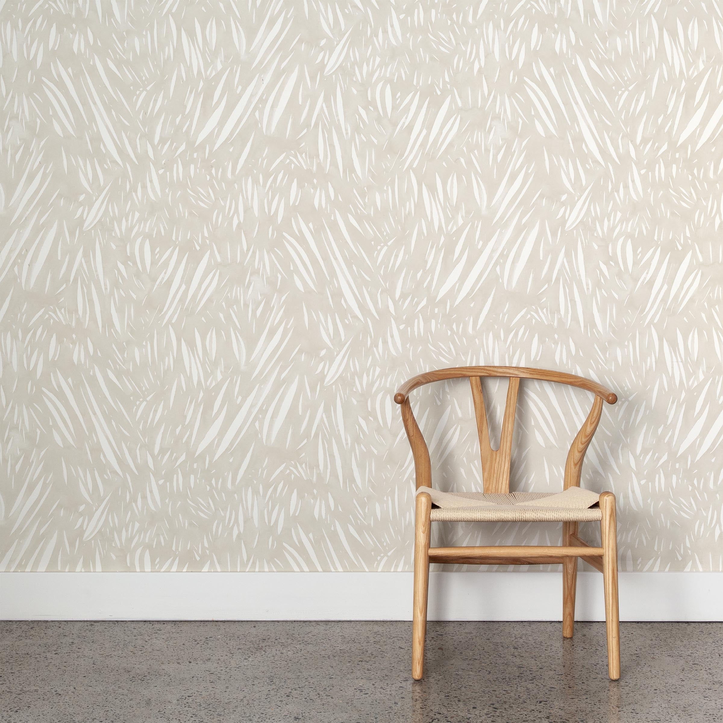 A wooden chair stands in front of a wall papered in an abstract leaf pattern in white on a soft beige watercolor field.