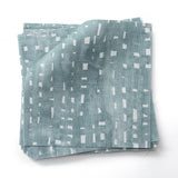 Stacked  fabric swatches in a painterly small-scale dashed dot print in white on a soft peacock blue watercolor field.