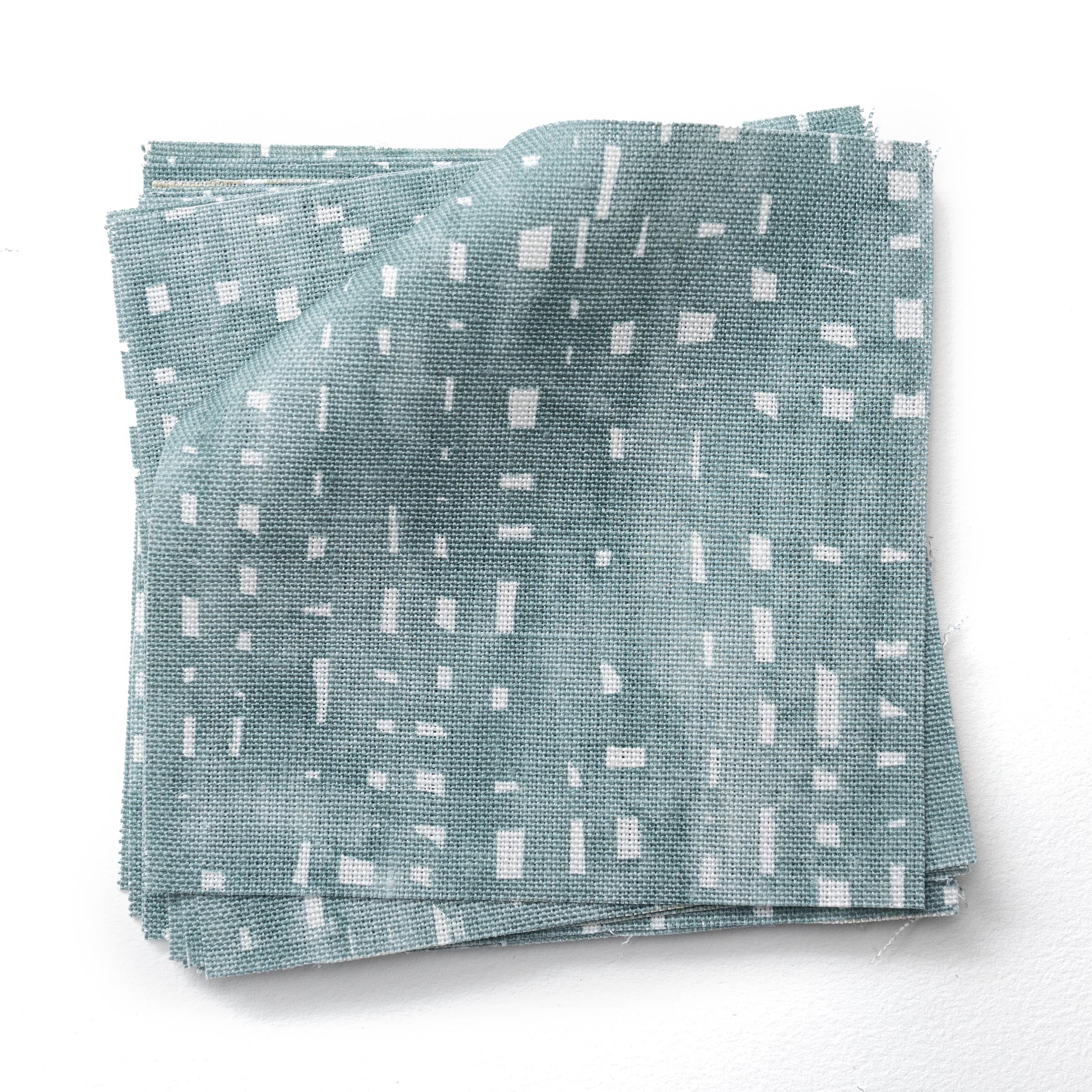 Stacked  fabric swatches in a painterly small-scale dashed dot print in white on a soft peacock blue watercolor field.