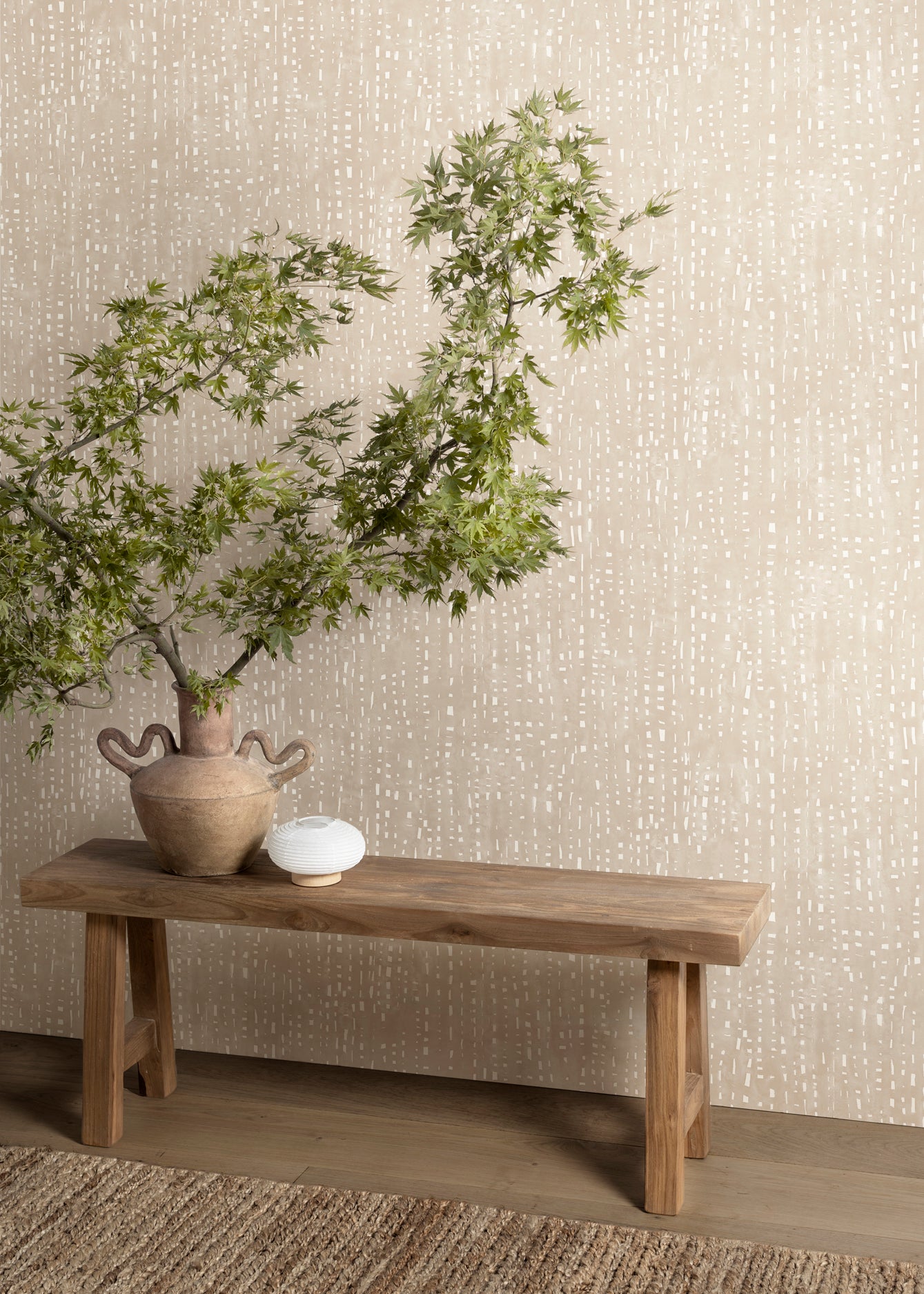 Wooden bench with urn and branches stands in front of a wall papered in a painterly dashed dot print in white on a soft beige field.