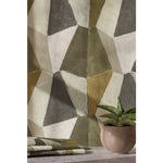 Detail image of a green patterned fabric and small spiky potted plant in front of a wall with mixed three dimensional diamond forms with black, white, taupe, dark forest green, dark sage green and a bright olive green on a textured paper, hover