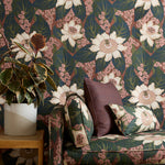 A styled interior corner featuring a moody pink and green floral wallpapered wall, a matching upholstered armchair with patterned cushions, a wooden side table, and a potted plant.