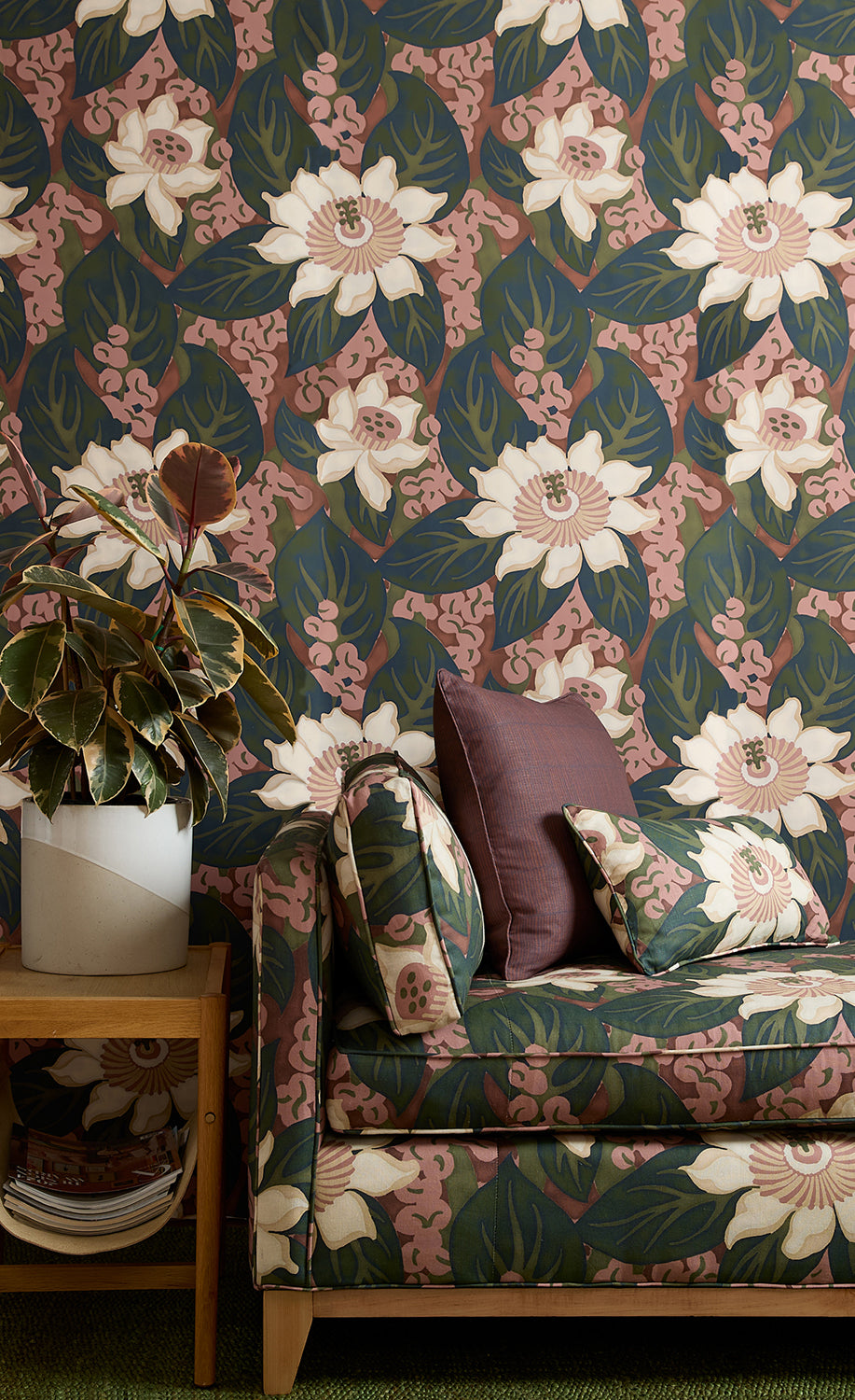 A styled interior corner featuring a moody pink and green floral wallpapered wall, a matching upholstered armchair with patterned cushions, a wooden side table, and a potted plant.