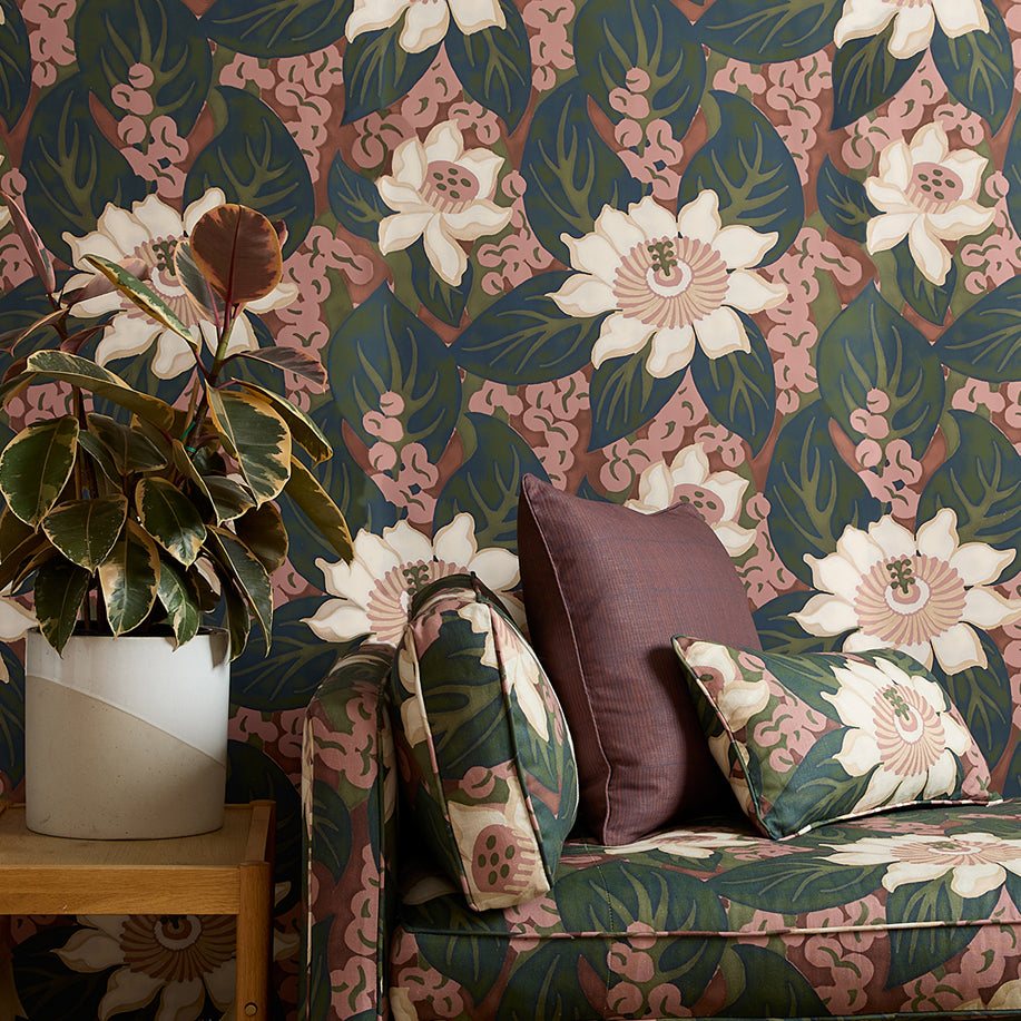 A styled interior corner featuring a moody pink and green floral wallpapered wall, a matching upholstered armchair with patterned cushions, a wooden side table, and a potted plant.