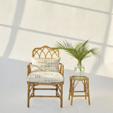 A rattan armchair with cream and tan mosaic cushions sits beside a small matching rattan stool holding a glass vase with a green palm frond, against a white wall with soft window-frame shadows.