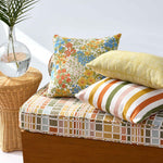 A styled outdoor daybed with a colorful mosaic-patterned cushion, a yellow striped pillow, and a floral patterned pillow, set next to a wicker side table with a palm leaf in a glass vase.