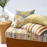 A styled outdoor daybed with a colorful mosaic-patterned cushion, a yellow striped pillow, and a floral patterned pillow, set next to a wicker side table with a palm leaf in a glass vase.