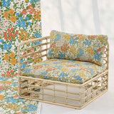 A rattan outdoor armchair with a cream-colored woven frame and floral cushions in blue, orange, yellow, and green on a cream background. A matching floral fabric panel is draped from the wall to the floor on the left, set against a bright white background.