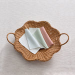 Three folded striped fabric swatches white, sage green, and dusty pink are arranged on a scalloped natural wicker tray against a textured off-white fabric background.