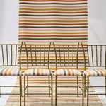 A styled studio scene showing a striped green, pink, yellow and white fabric backdrop extending from the wall to the floor, with four green bamboo chairs with matching seat cushions. 