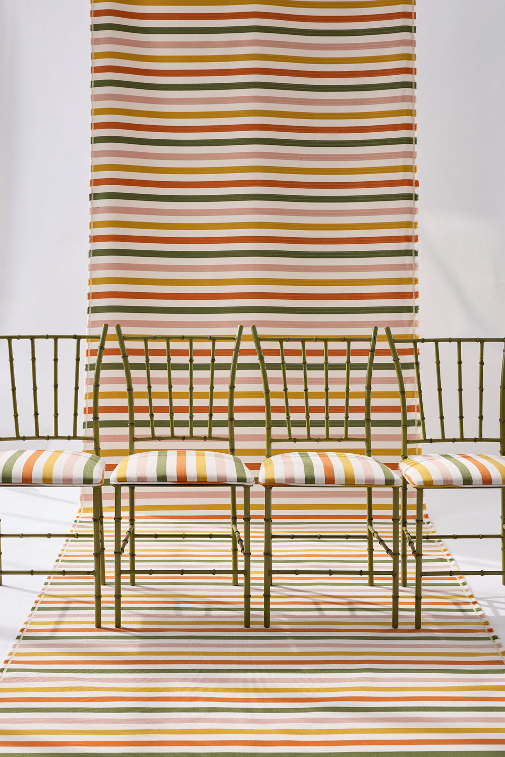 A styled studio scene showing a striped green, pink, yellow and white fabric backdrop extending from the wall to the floor, with four green bamboo chairs with matching seat cushions. 