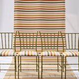 A styled studio scene showing a striped green, pink, yellow and white fabric backdrop extending from the wall to the floor, with four green bamboo chairs with matching seat cushions. 
