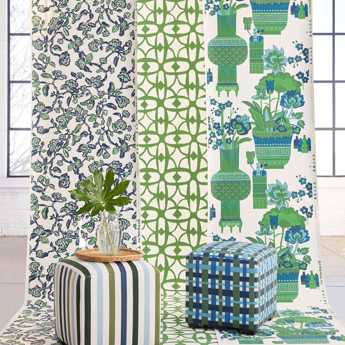 Three vertical panels of patterned wallpaper displayed in a bright room, with two upholstered ottomans in front. The left panel has a navy and green floral pattern on white; the center panel has a green geometric lattice pattern on white; the right panel features blue and green floral motifs with potted plants on white.