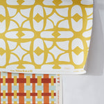 Close up of two wallpapers, the top is a yellow and white lattice design layered over a basketweave plaid in red, orange and yellow. 