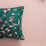 A square decorative throw pillow with a dark teal green background and a black, white, and dusty pink floral pattern, shown against a dusty pink lattice patterned wallpaper.
