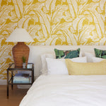 A styled bedroom featuring a bold yellow and white tropical leaf patterned wallpaper on the wall behind a bed with white bedding, decorative pillows, a wicker bedside table, and a lamp.