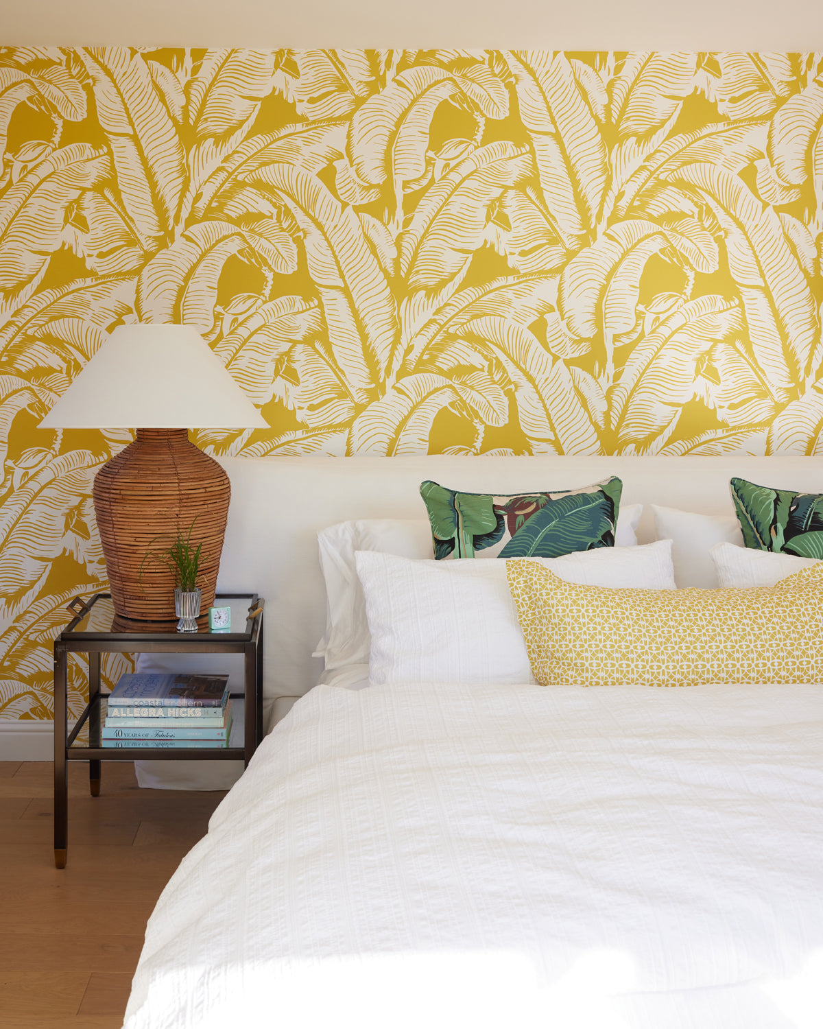 A styled bedroom featuring a bold yellow and white tropical leaf patterned wallpaper on the wall behind a bed with white bedding, decorative pillows, a wicker bedside table, and a lamp.