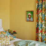 A cozy bedroom corner featuring mustard yellow patterned wallpaper, a bed with white bedding and colorful floral pillows, a mint green throw blanket, and a matching floral curtain.