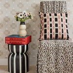 A high-back dining chair with a beige and black leopard print slipcover, with a throw pillow in pink, tan and black plaid, with a black and white striped side table with red books, and cream ceramic vase with flowers against a beige floral lattice wallpaper. 