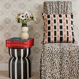 A high-back dining chair with a beige and black leopard print slipcover, with a throw pillow in pink, tan and black plaid, with a black and white striped side table with red books, and cream ceramic vase with flowers against a beige floral lattice wallpaper. 