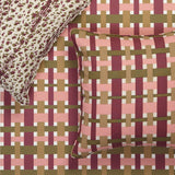 A close-up of a woven-texture throw pillow and matching pillowcase set on a bed. The pillow features a pink, burgundy, and olive green basketweave pattern, while the matching pillowcase has a leopard print design in cream and burgundy.