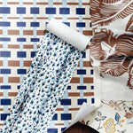 A styled flat-lay of patterned wallpaper sheets, featuring a central roll of white wallpaper with blue and black abstract spots, alongside other sheets with geometric brown and blue patterns and a floral print.