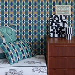 A styled bedroom corner featuring a bold geometric wallpaper in teal, navy, and beige, a wooden nightstand, a striped lamp, and patterned pillows on a bed.