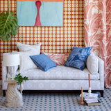 A styled living room scene featuring a light beige upholstered sofa with patterned blue and coral pillows, set against a bold orange and yellow checkered wallpaper wall. A matching coral and cream palm-patterned curtain hangs on the right, and a white side table with a lamp sits on the left.