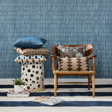 A styled interior scene with a wooden armchair holding patterned throw pillows, a matching patterned pillow stacked on a white cylindrical stool, and a blue textured wallpaper behind them.
