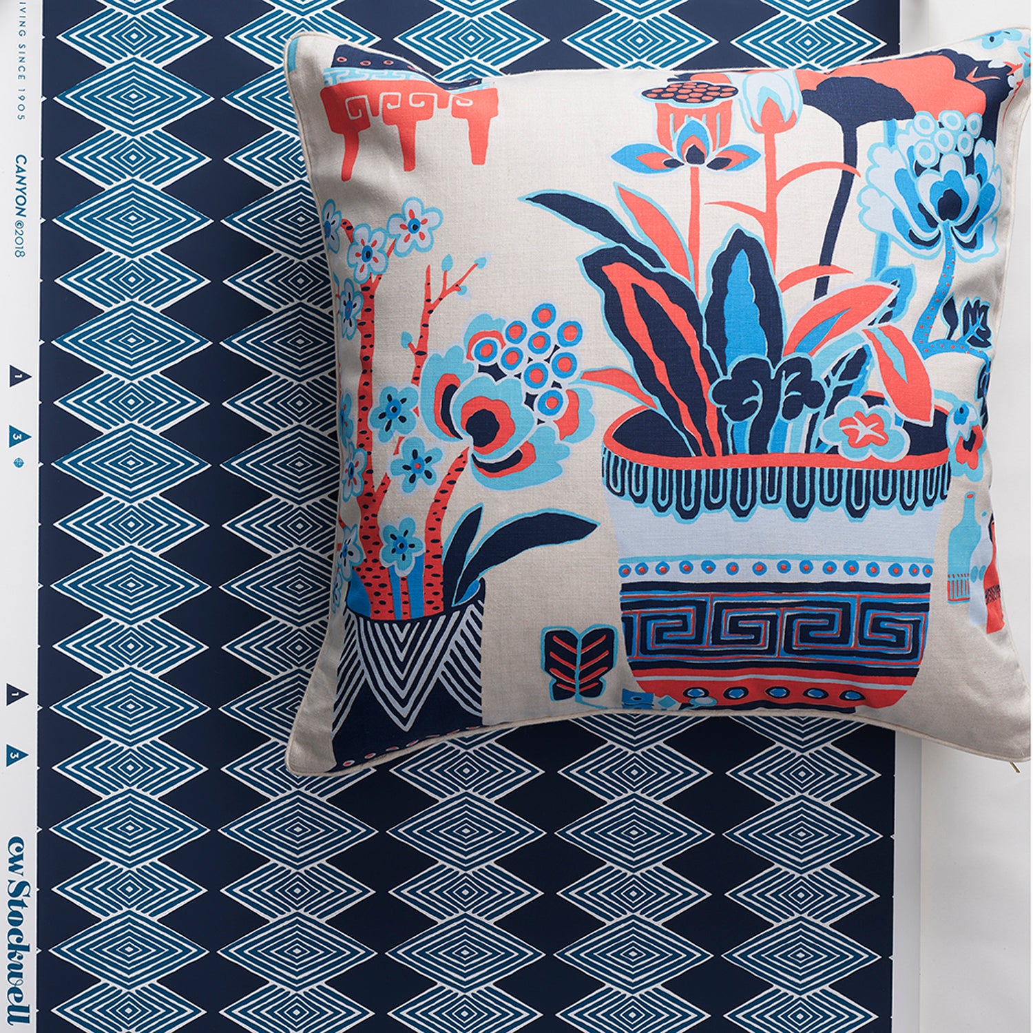 A square throw pillow with a colorful botanical and vase pattern, displayed against a navy blue diamond stripe wallpaper backdrop. The pillow features a cream base with blue, coral red, and navy floral motifs.