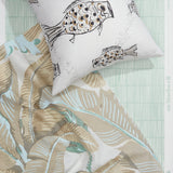 A styled flat-lay with banana leaf pattered fabric in tan and seafoam, with throw pillow with black and white fish pattern atop a wallpaper with a bamboo stripe pattern in aqua blue and white. 