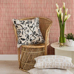 Living room scene with wicker chair with a neutral color floral branch pattern, white side table with small potted plant, glass vase with white callayilies and  brass elephant with stacked neutral color patterned throw pillows in the foreground. 