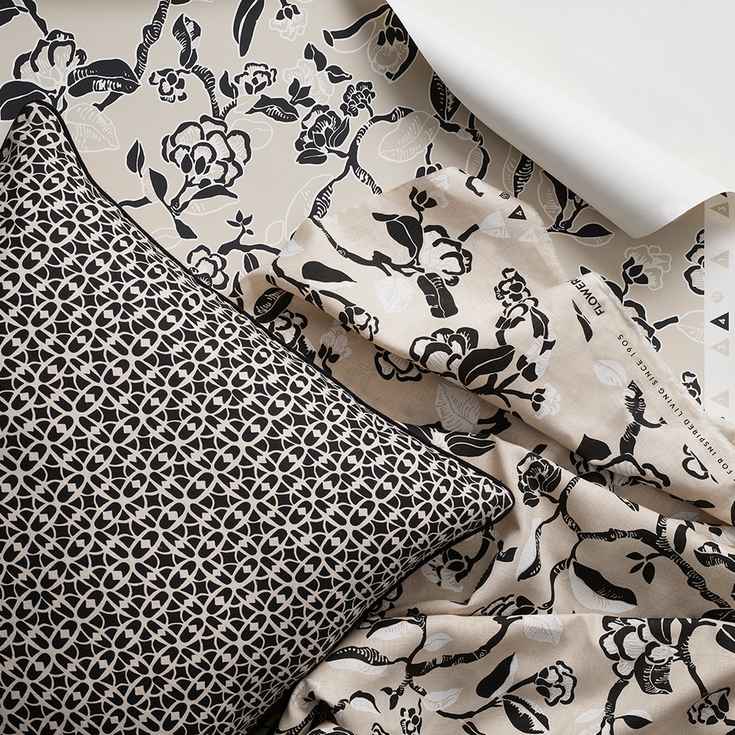 A styled flat-lay image with a throw pillow in a small scale black and white lattice design layered over a hand drawn branch and flower fabric and wallpaper in black, white and beige. 
