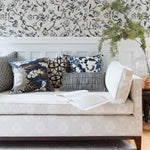 A styled living room scene featuring a cream upholstered daybed with multiple decorative throw pillows, set against a white wainscoted wall with black floral wallpaper and a glass vase with greenery.