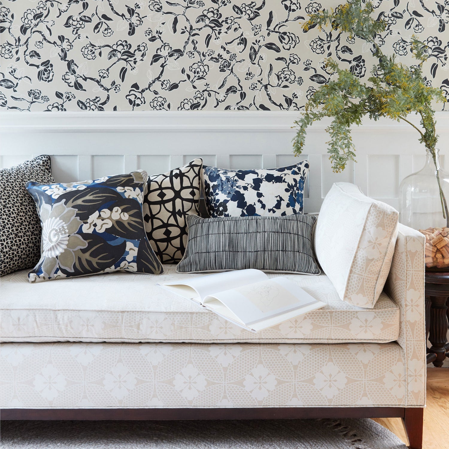 A styled living room scene featuring a cream upholstered daybed with multiple decorative throw pillows, set against a white wainscoted wall with black floral wallpaper and a glass vase with greenery.