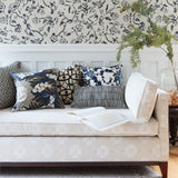 A styled living room scene featuring a cream upholstered daybed with multiple decorative throw pillows, set against a white wainscoted wall with black floral wallpaper and a glass vase with greenery.