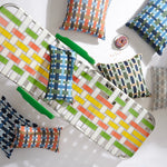 A woven metal lounge chair with a colorful geometric basketweave pattern and green armrests, styled with several matching patterned throw pillows on a white background.
