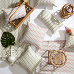 A styled flat-lay of patterned throw pillows and matching textiles arranged on a white surface with wicker and rattan decor. The textiles and pillows feature a small repeating geometric motif in muted green and warm orange/terracotta on an off-white base.