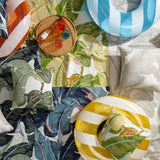 Styled flat-lay image featuring bold banana leaf fabrics in an arrange of bright colors layered with striped pool tubes and a wicker tray with bottle of rose wine and colorful glasses. 