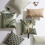 A styled flat-lay of multiple decorative throw pillows in different patterns and colors, arranged on a white background with a vintage tennis racket, sunglasses, and a small green bottle as props.